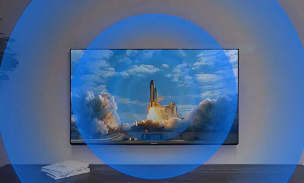 Telewizor wyświetla obraz startującej rakiety na tle nieba z chmurami. Wokół ekranu widoczny jest niebieski okrąg, a na półce poniżej leży biały przedmiot.