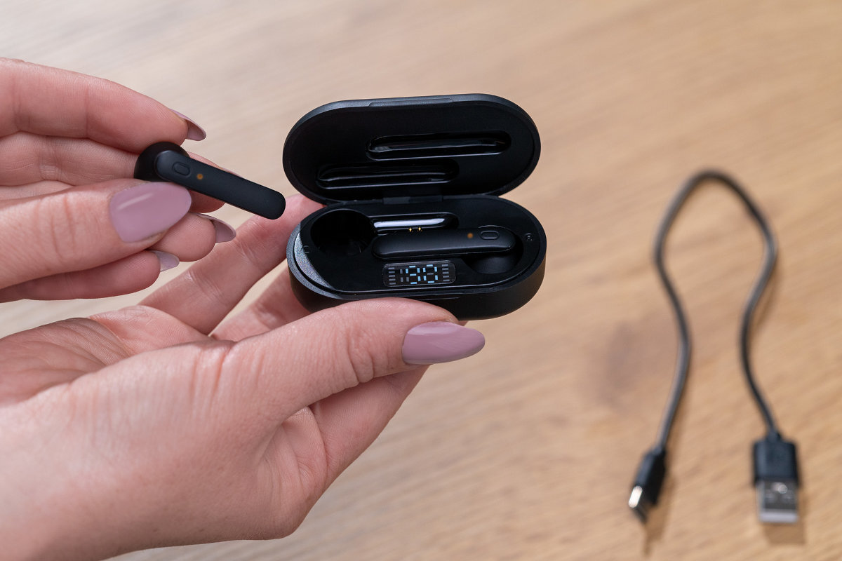 Osoba trzyma czarne słuchawki douszne oraz otwarte etui ładujące z wyświetlaczem. Obok leży krótki kabel USB.