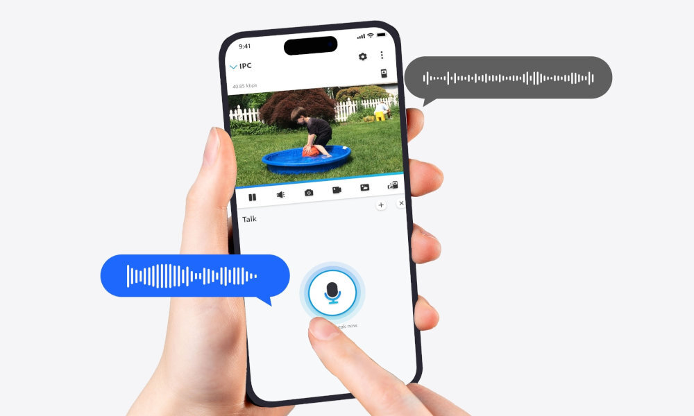 Osoba trzyma smartfon, na którym wyświetlany jest obraz dziecka bawiącego się w basenie. Na ekranie widoczne są ikony aplikacji oraz niebieska ikona mikrofonu z napisem \'Hold to talk\'.