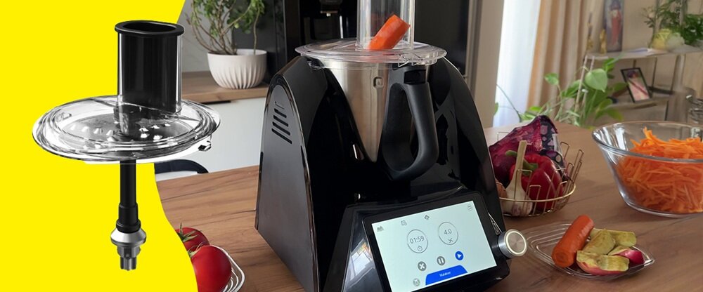Na blacie kuchennym stoi czarny robot kuchenny z wyświetlaczem dotykowym, w którym znajduje się marchewka. Obok widoczna jest przezroczysta pokrywa z elementem do przetwarzania żywności oraz miska z pokrojonymi warzywami.