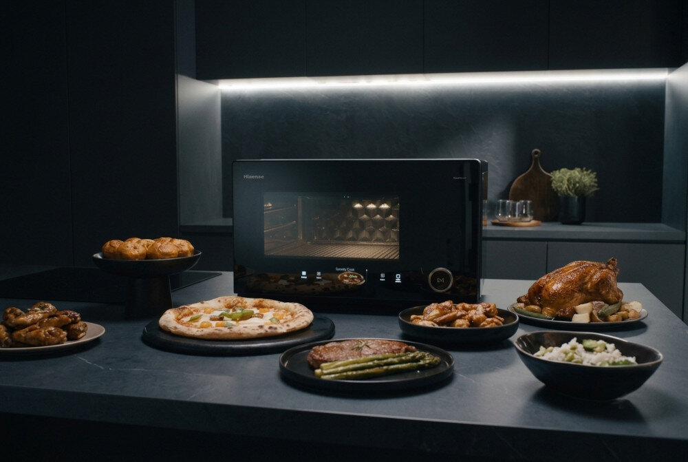 Na blacie kuchennym stoi kuchenka mikrofalowa marki Hisense, otoczona różnorodnymi potrawami na talerzach, w tym pizza, pieczony kurczak i warzywa. W tle widoczna jest nowoczesna kuchnia z oświetleniem podszafkowym.