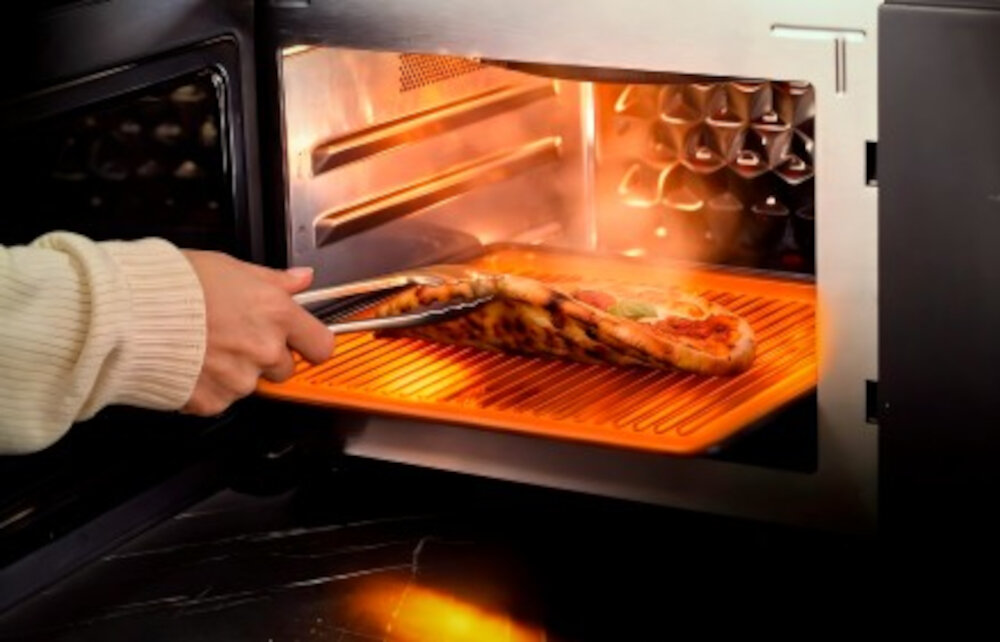 Osoba wyciąga pizzę z kuchenki mikrofalowej za pomocą szczypiec. Wnętrze kuchenki jest oświetlone, a na dolnej półce znajduje się pomarańczowa tacka.