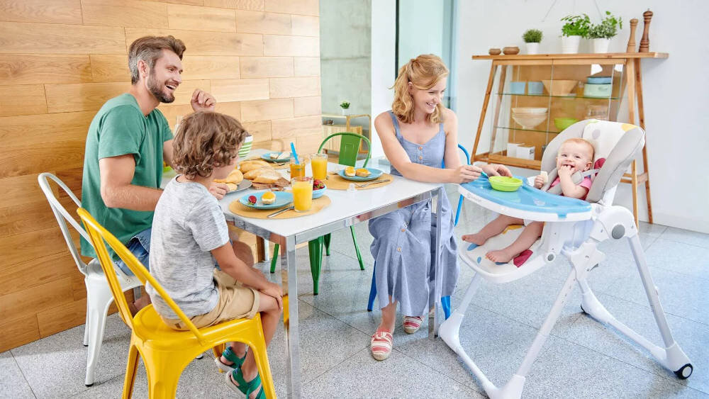 Rodzina siedzi przy stole, na którym znajdują się talerze z jedzeniem i szklanki z napojami. Dziecko siedzi w szarym krzesełku do karmienia KINDERKRAFT Yummy.