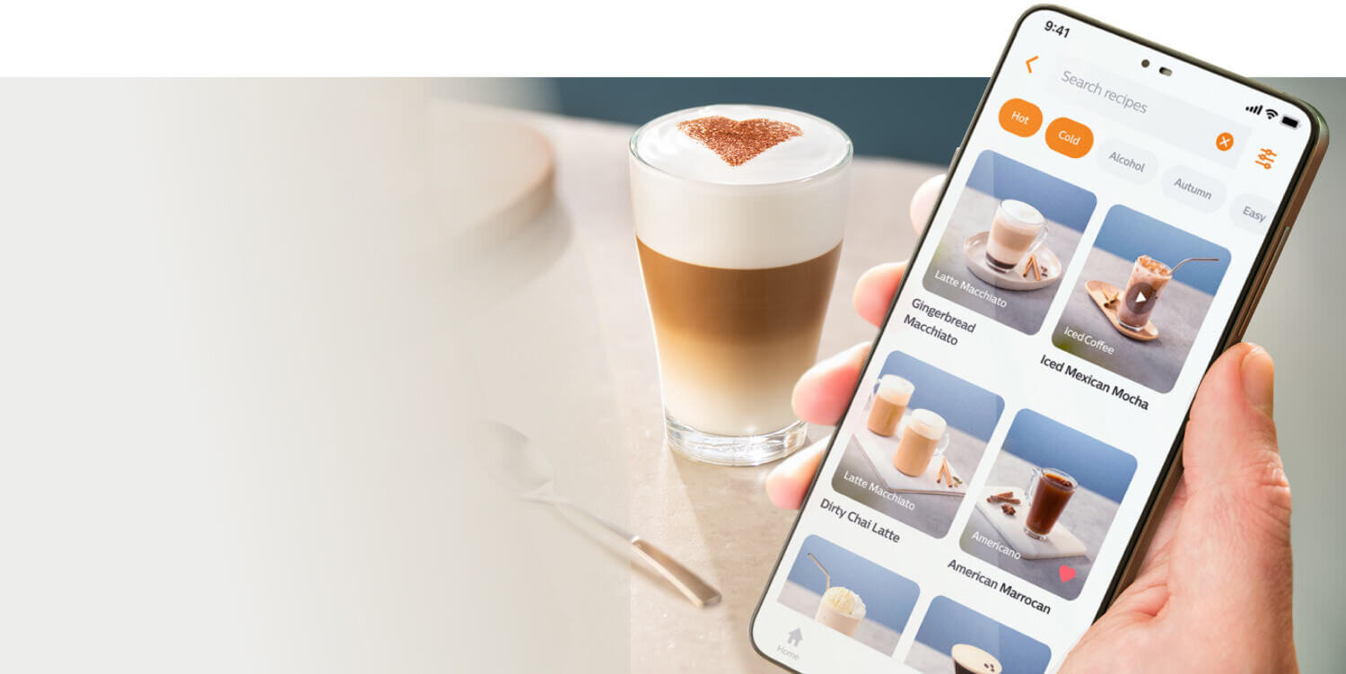 Osoba trzyma smartfon z aplikacją do wyboru przepisów na kawę, obok stoi szklanka z kawą z pianką i wzorem na wierzchu. Na ekranie telefonu widoczne są różne rodzaje kaw, takie jak \'Gingerbread Macchiato\' i \'Iced Mexican Mocha\'.