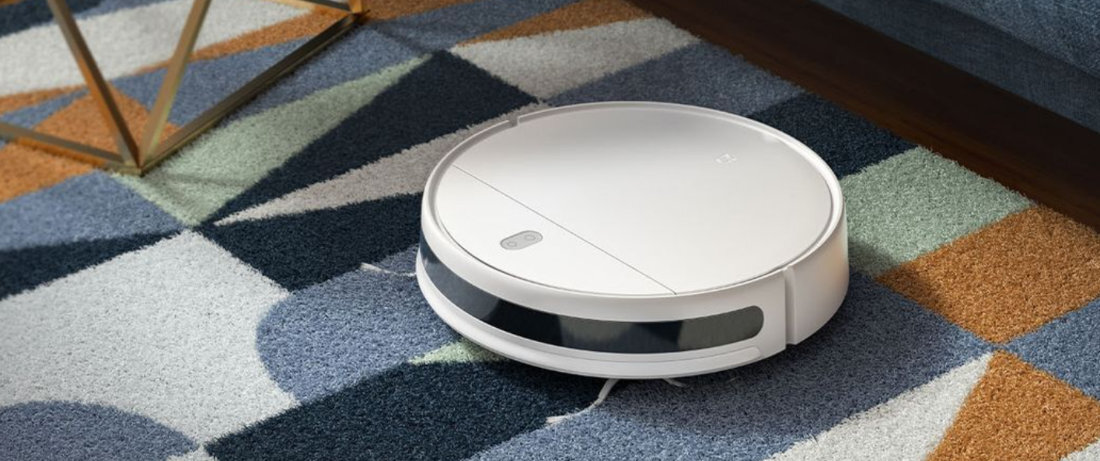 Na kolorowym dywanie znajduje się biały robot sprzątający o okrągłym kształcie. Robot ma ciemny panel z przodu i jest włączony.