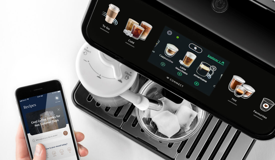 Na obrazie widoczny jest ekspres do kawy z wyświetlaczem pokazującym różne rodzaje napojów, takie jak cappuccino i latte macchiato. Obok ekspresu znajduje się telefon z aplikacją wyświetlającą przepis na kawę na gorące letnie dni.