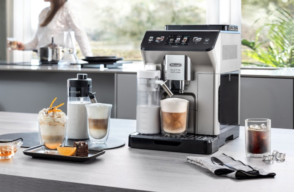 Na blacie kuchennym stoi ekspres do kawy z logo \'DeLonghi Eletta Explore\', obok którego znajdują się różne napoje kawowe i deser. W tle widać osobę przy oknie oraz inne elementy kuchenne.