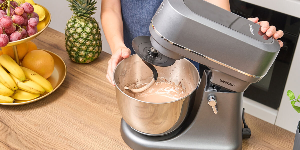 Osoba obsługuje szary robot kuchenny z misą, w której znajduje się mieszana masa. W tle widoczne są owoce, takie jak ananas i banany, a na urządzeniu znajduje się logo \'Zelmer\'.