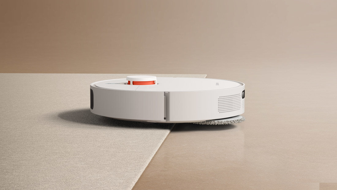Na obrazie widoczny jest biały robot sprzątający poruszający się z powierzchni dywanu na gładką podłogę. Robot ma okrągły kształt i widoczny jest pomarańczowy element na górze.