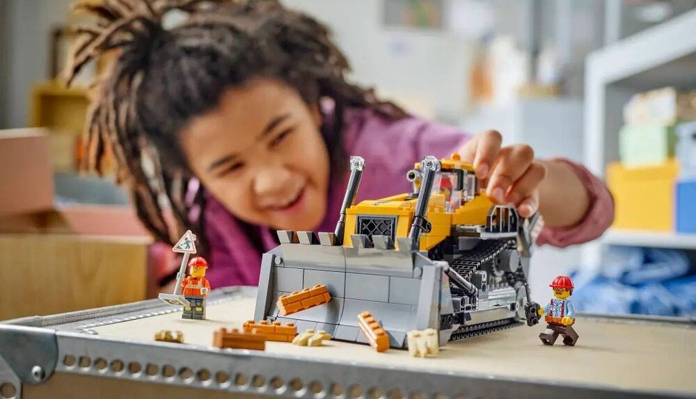 Na obrazie widać dziecko bawiące się modelem żółtego buldożera z klocków LEGO. Obok buldożera znajdują się dwie figurki robotników i kilka elementów przypominających cegły.