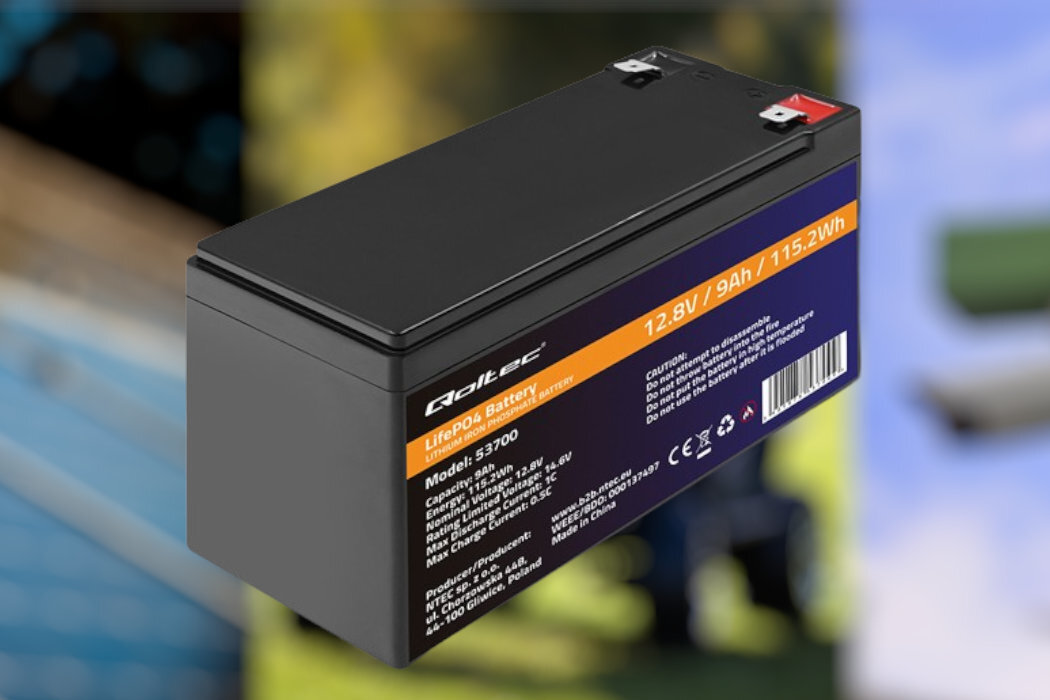 Na obrazie widoczny jest czarny akumulator z oznaczeniami: \'Qoltec\', \'LiFePo4 Battery\', \'Model: 53700\', \'12.8V / 9Ah / 115.2Wh\'. W tle rozmyte elementy, które nie są istotne dla opisu produktu.