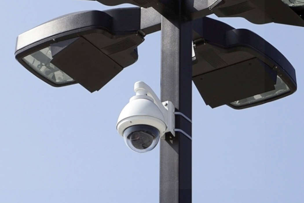 Na obrazie widoczna jest kamera monitoringu zamontowana na słupie, obok dwóch lamp ulicznych. Tło stanowi jasne, bezchmurne niebo.