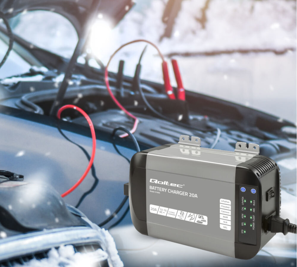 Na obrazie widoczna jest inteligentna ładowarka Qoltec Battery Charger 20A z kilkoma ikonami i wskaźnikami na obudowie. W tle znajduje się otwarta maska samochodu z podłączonymi kablami rozruchowymi.