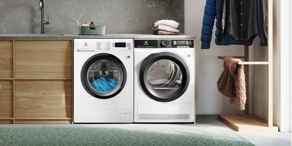 Na obrazie znajduje się pralka i suszarka marki ELECTROLUX ustawione obok siebie w nowoczesnej pralni. Obok urządzeń znajduje się drewniana szafka, a na niej koszyk z detergentami.
