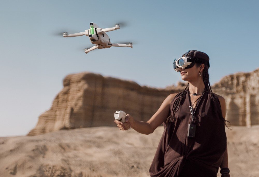 Osoba w okularach VR steruje dronem unoszącym się w powietrzu na tle skalistego krajobrazu. Dron jest biały z zielonymi elementami.