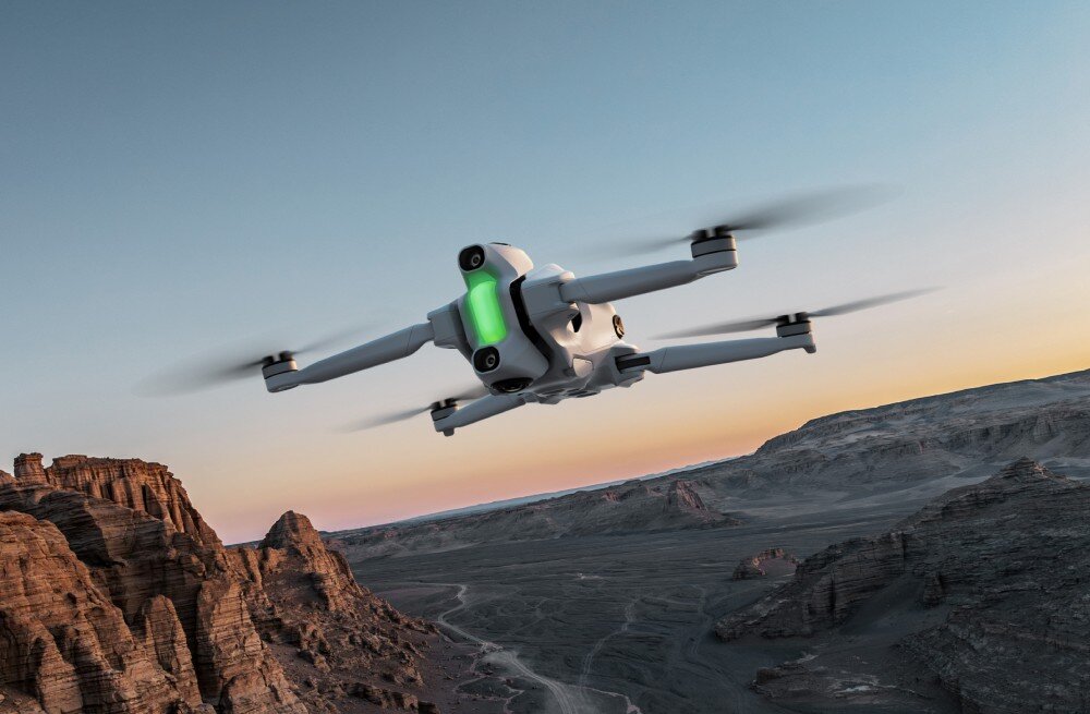 Dron z zielonym światłem na korpusie unosi się nad skalistym krajobrazem o zachodzie słońca. W tle widać formacje skalne i rozległą dolinę.