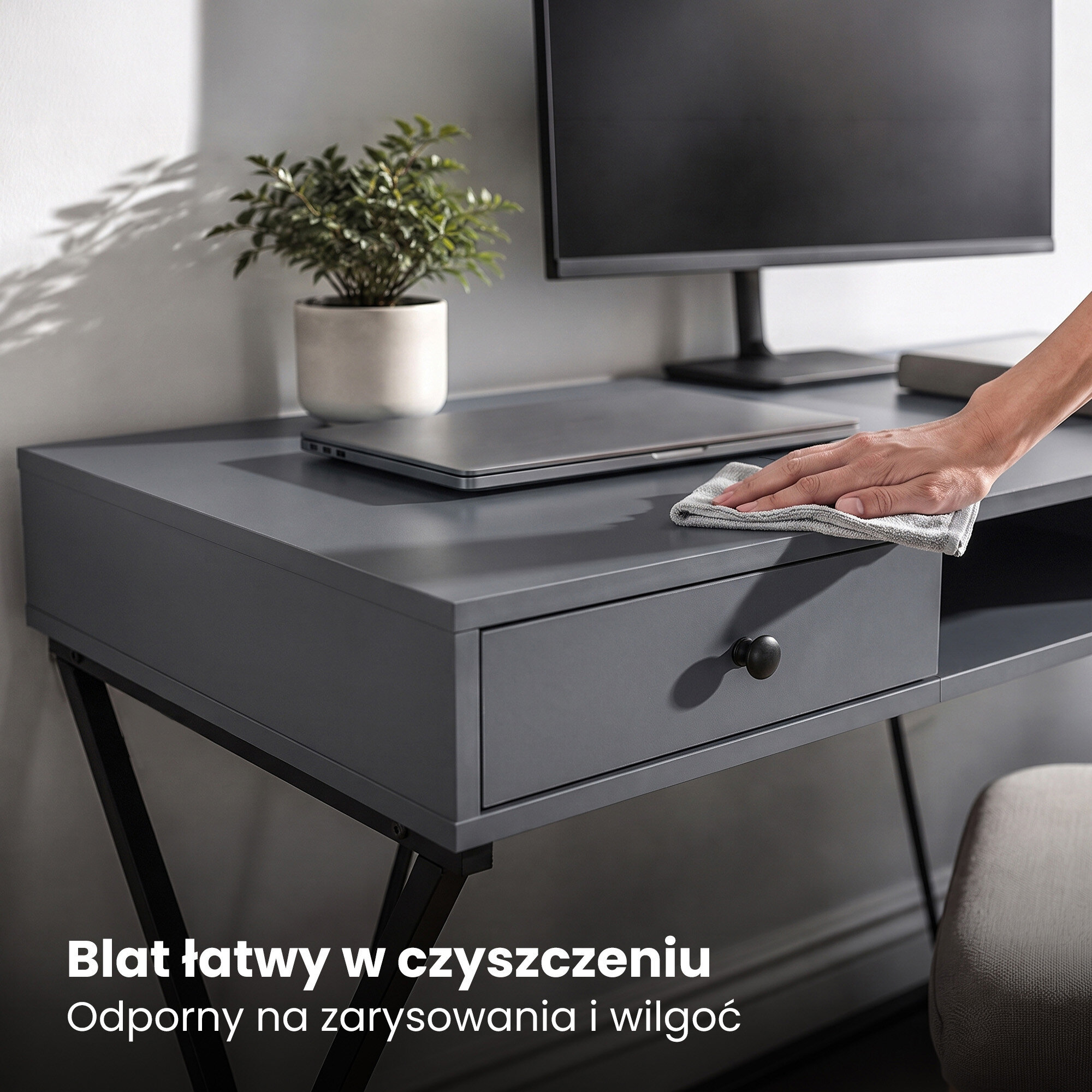 Na obrazie widoczne jest grafitowe biurko z czarnymi nogami, na którym stoi monitor i laptop. Na blacie znajduje się tekst: \'Blat łatwy w czyszczeniu Odporny na zarysowania i wilgoć\'.
