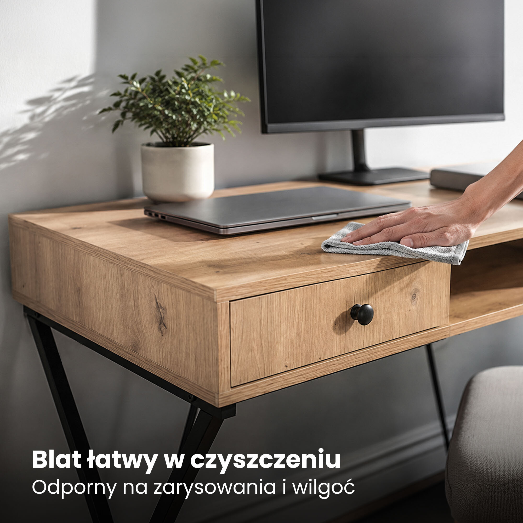 Drewniane biurko z czarnymi nogami, na którym znajduje się laptop, monitor i roślina w doniczce. Na blacie widoczna jest ręka czyszcząca powierzchnię ściereczką, a na dole napis: \'Blat łatwy w czyszczeniu. Odporny na zarysowania i wilgoć\'.