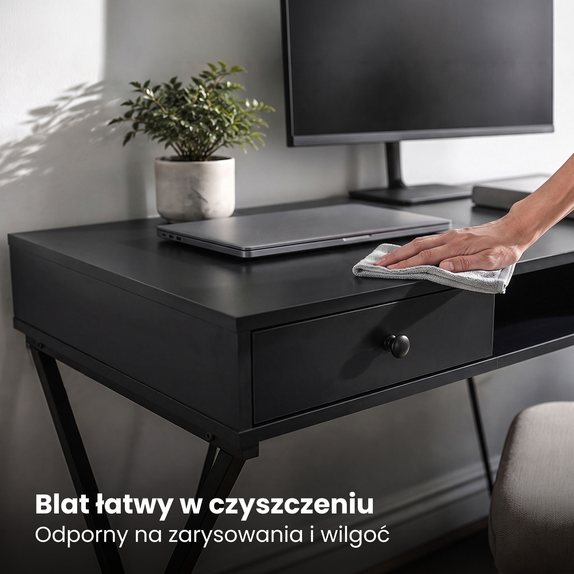 Czarne biurko z dwoma szufladami i jedną półką, na którym znajduje się laptop, monitor oraz roślina w doniczce. Na blacie widoczna jest ręka czyszcząca powierzchnię ściereczką, a na dole zdjęcia tekst: \'Blat łatwy w czyszczeniu. Odporny na zarysowania i wilgoć\'.