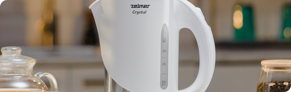 Na obrazie widoczny jest biały czajnik elektryczny z przezroczystym wskaźnikiem poziomu wody. Na czajniku znajduje się logo \'zelmer Crystal\'.