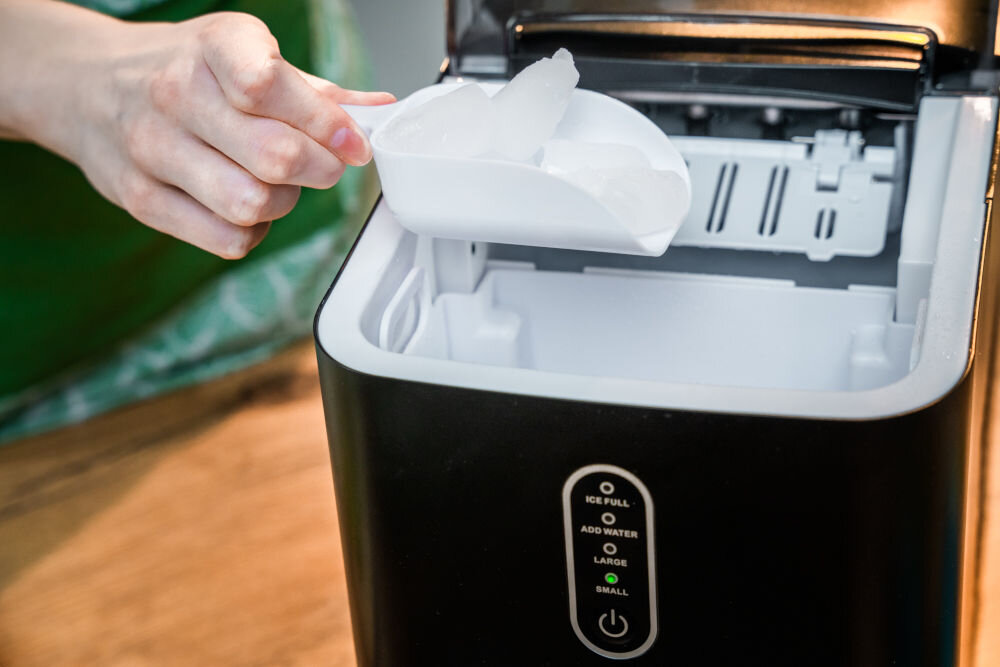 Osoba trzyma łopatkę z lodem nad otwartą kostkarką do lodu. Na urządzeniu widoczne są kontrolki z napisami: \'ICE FULL\', \'ADD WATER\', \'LARGE\', \'SMALL\'.