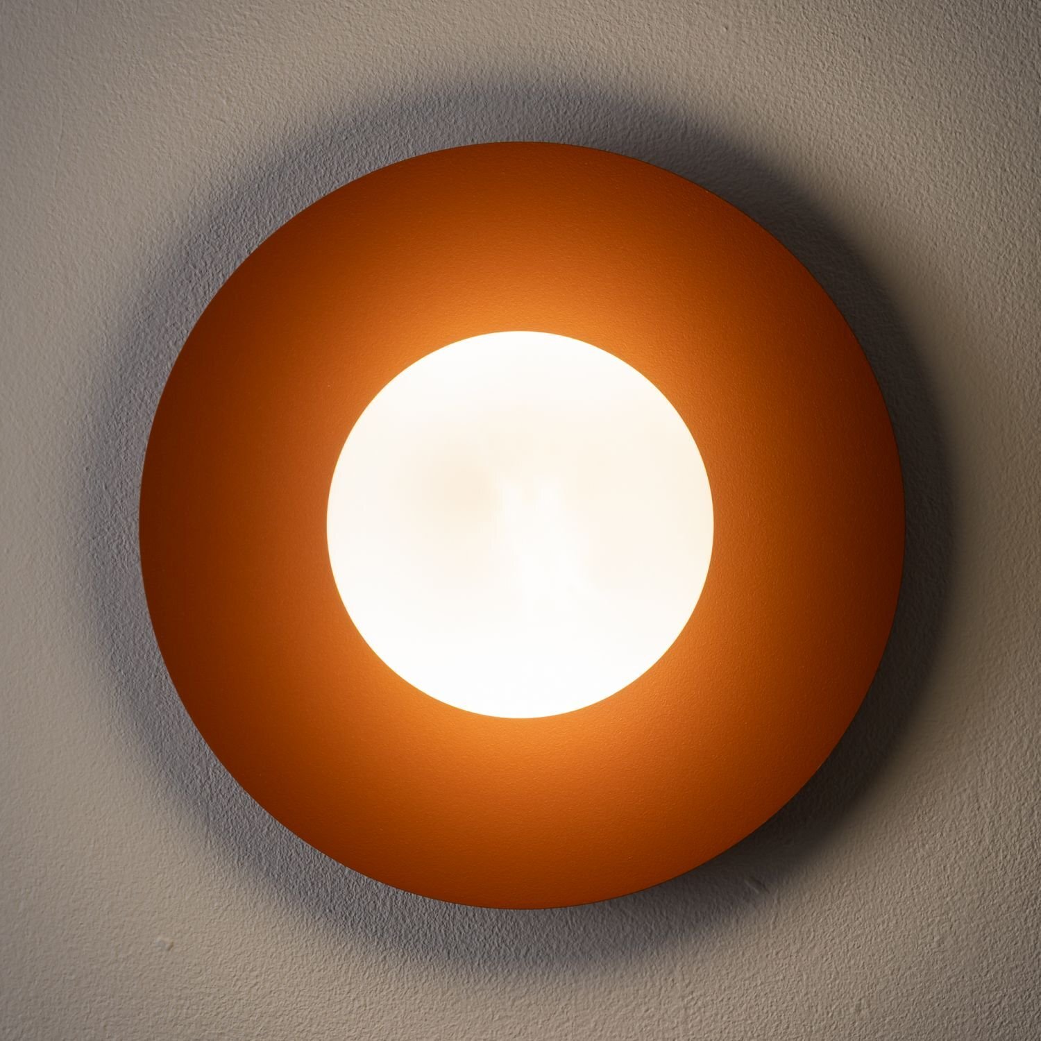 Na obrazie widoczna jest okrągła lampa ścienna z jasnym, centralnym źródłem światła otoczonym pomarańczowym pierścieniem. Lampa jest zamontowana na ścianie.