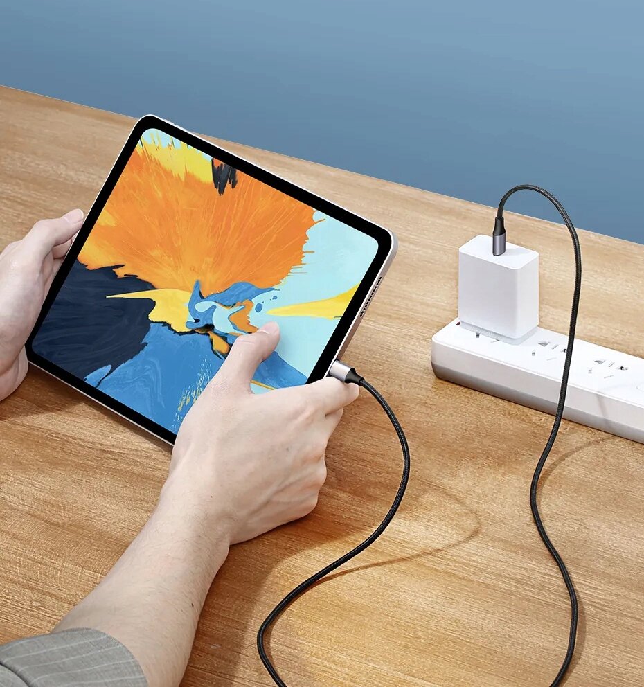 Osoba trzyma tablet z kolorowym ekranem, podłączony do ładowarki za pomocą czarnego kabla USB-C. Ładowarka jest wpięta do listwy zasilającej na drewnianym stole.