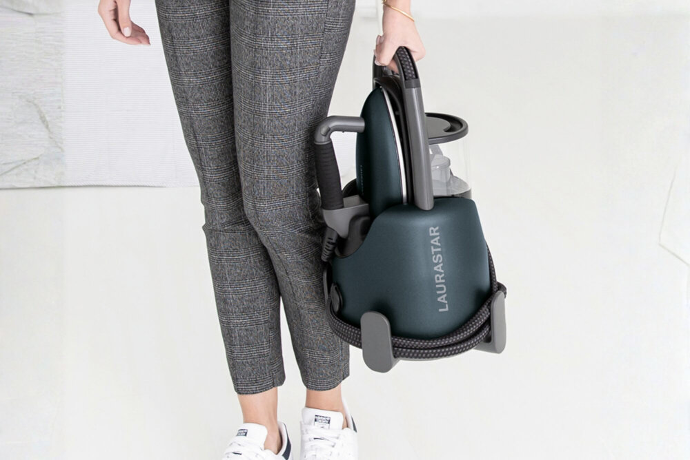 Osoba trzyma generator pary w kolorze ciemnoniebieskim z widocznym napisem \'LAURASTAR\'. Urządzenie ma ergonomiczny uchwyt i kompaktowy design.
