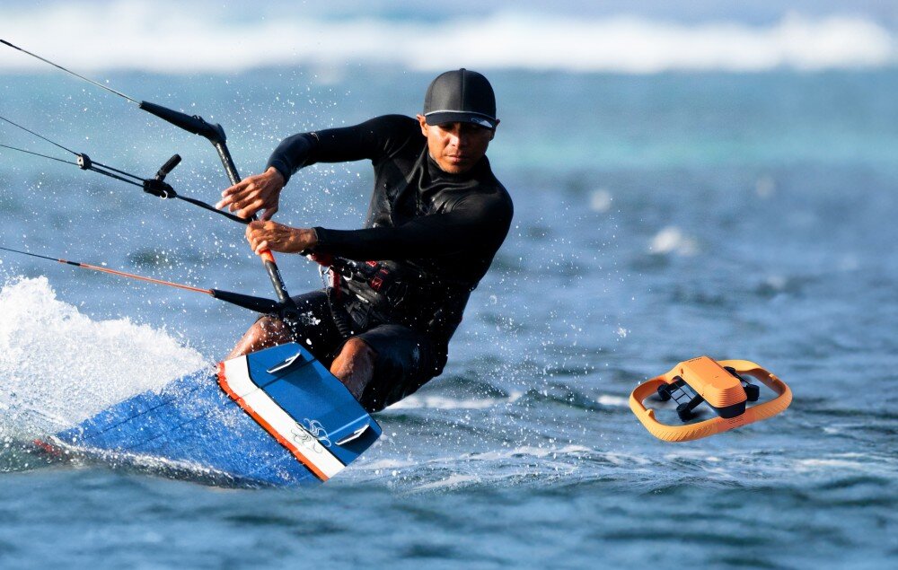 Osoba uprawiająca kitesurfing na wodzie, obok niej unosi się pomarańczowy dron wodny. W tle widać rozmytą linię horyzontu z falami.