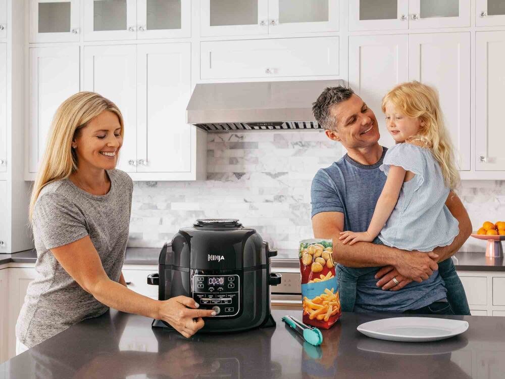 Na blacie kuchennym stoi multicooker marki Ninja, obok którego znajduje się kobieta obsługująca urządzenie. W tle widać mężczyznę trzymającego dziecko na rękach oraz pojemnik z frytkami.