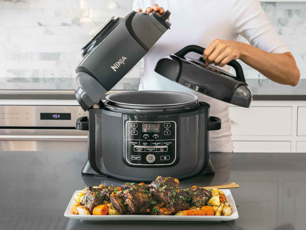 Osoba obsługuje multicooker marki Ninja, który jest otwarty i znajduje się na blacie kuchennym. Przed urządzeniem leży talerz z gotowym daniem mięsnym i warzywami.