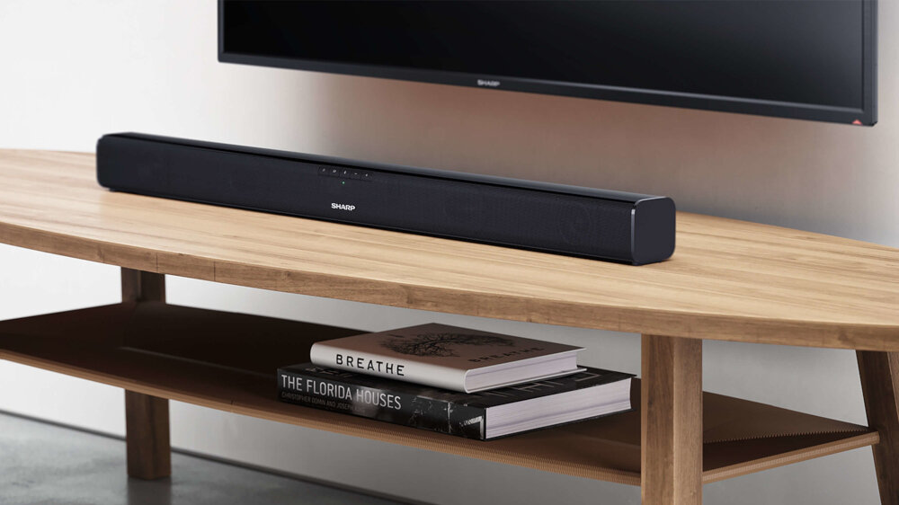 Czarny soundbar marki SHARP umieszczony na drewnianej półce pod telewizorem. Na półce znajdują się również dwie książki z widocznymi tytułami: \'BREATHE\' i \'THE FLORIDA HOUSES\'.