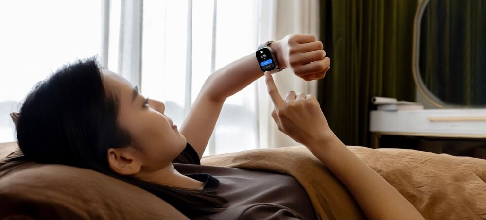 Osoba leży na łóżku i patrzy na smartwatch na nadgarstku, dotykając jego ekranu. W tle widoczne są zasłony i toaletka.