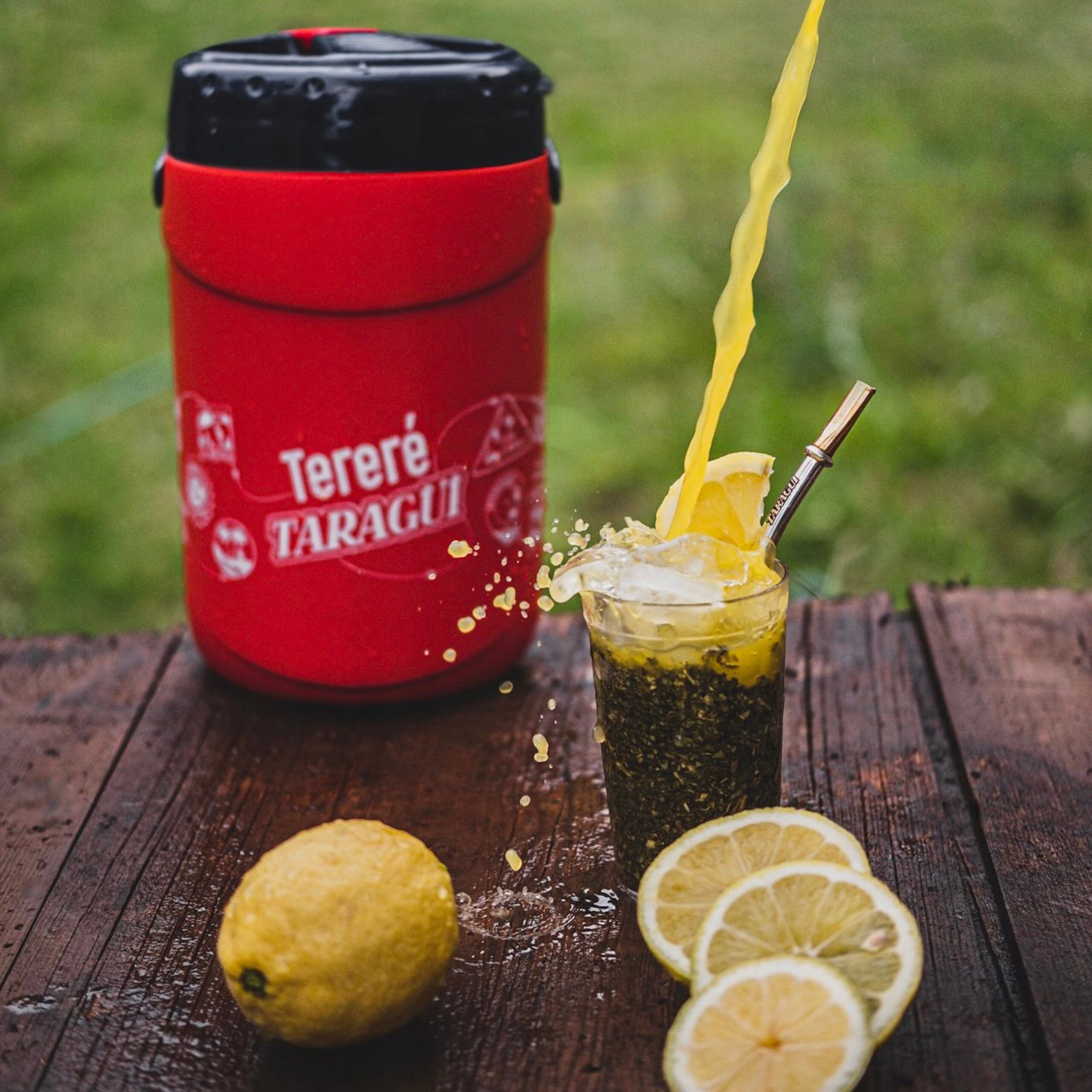 Na drewnianym stole stoi czerwony termos z napisem \'Tereré Taragui\'. Obok znajduje się szklanka z yerba mate, plasterkami cytryny i metalową rurką, a także całe i pokrojone cytryny.