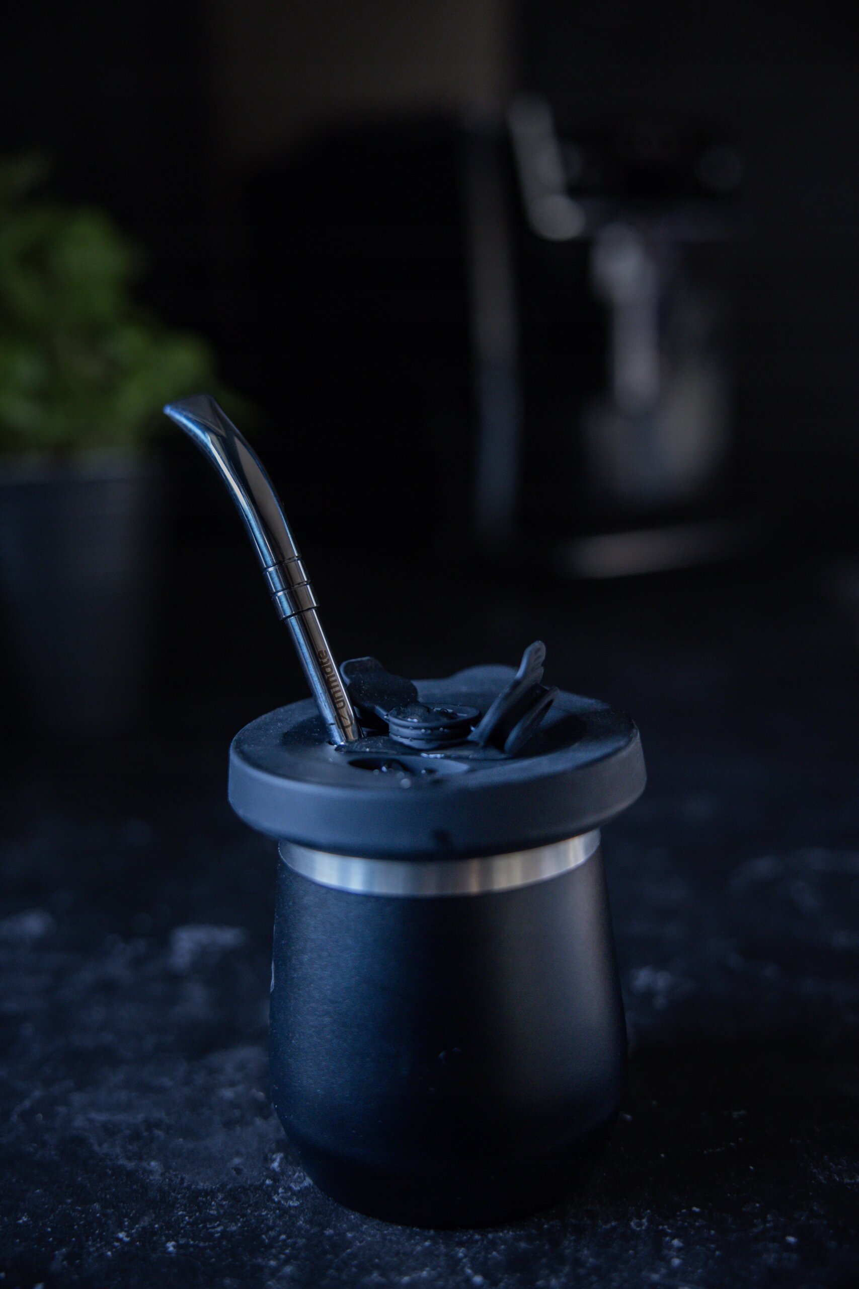 Na ciemnym tle znajduje się czarny kubek z metalową rurką do picia yerba mate. W tle widoczna jest rozmyta sylwetka rośliny i urządzenia.