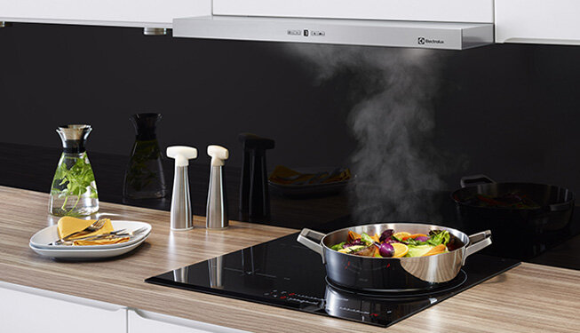 Na blacie kuchennym znajduje się płyta indukcyjna z gotującym się jedzeniem w garnku, a nad nią zamontowany jest okap z logo Electrolux. Obok płyty leży talerz z jedzeniem oraz karafka z wodą i ziołami.