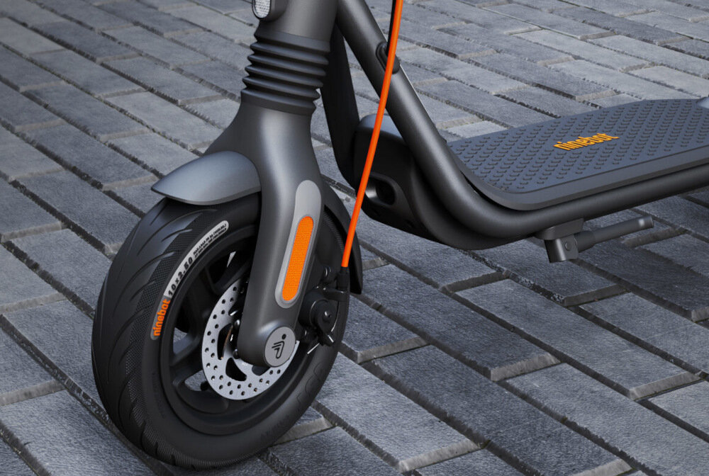 Zbliżenie na przednie koło czarnej hulajnogi elektrycznej z widocznym amortyzatorem i tarczą hamulcową. Na oponie widoczny jest napis \'tubeless\'.
