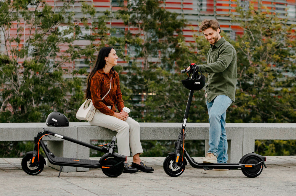 Dwie osoby rozmawiają na zewnątrz, obok nich znajdują się dwie czarne hulajnogi elektryczne. Na jednej z hulajnóg widoczne jest logo \'Segway\'.