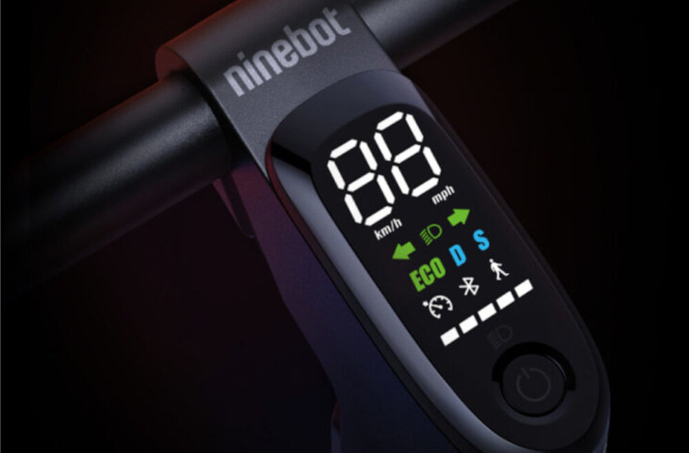 Wyświetlacz hulajnogi elektrycznej pokazuje prędkość w km/h i mph oraz tryby ECO, D i S. Widoczne są również ikony świateł, Bluetooth, tempomatu i poziomu baterii, a na górze znajduje się napis \'ninebot\'.