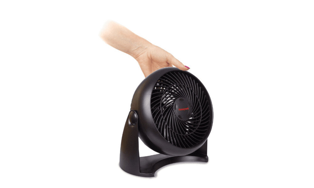 Czarny wentylator z widocznym logo \'Honeywell\' na środku, trzymany przez rękę. Wentylator ma okrągłą obudowę i stoi na podstawce.