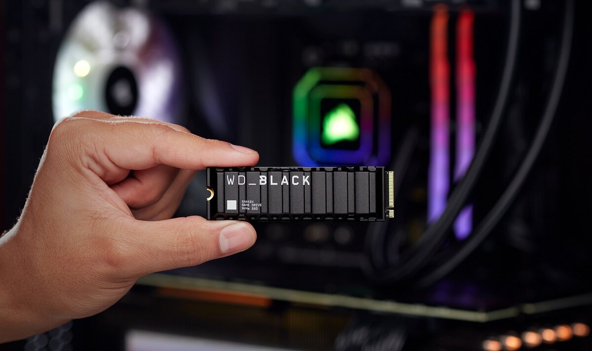 Osoba trzyma dysk SSD z napisem \'WD_BLACK\' na tle wnętrza komputera z podświetleniem RGB.
