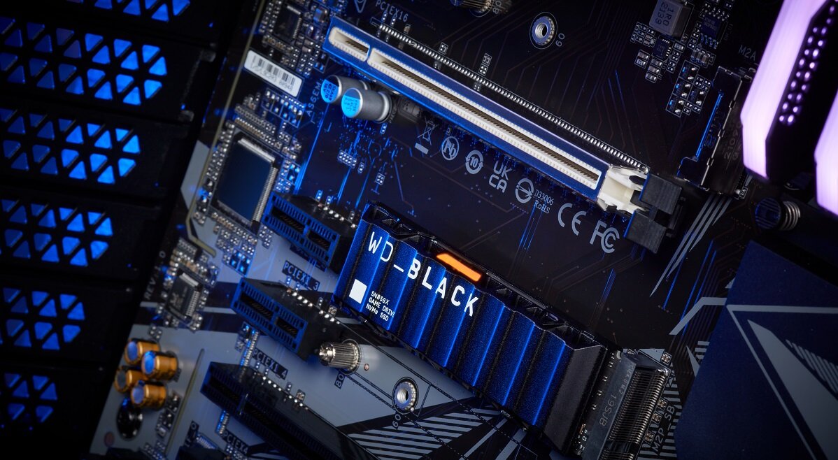 Na obrazie widoczna jest płyta główna komputera z zamontowanym dyskiem SSD oznaczonym jako WD_BLACK. W tle widać elementy elektroniczne i złącza, a całość jest oświetlona niebieskim światłem.