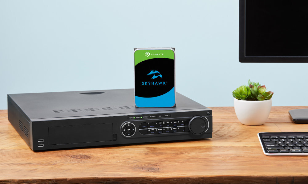 Na drewnianym biurku znajduje się czarny sprzęt elektroniczny z panelem kontrolnym oraz dysk z napisem \'SEAGATE SkyHawk AI\'. Obok stoi mała doniczka z zieloną rośliną i fragment klawiatury.