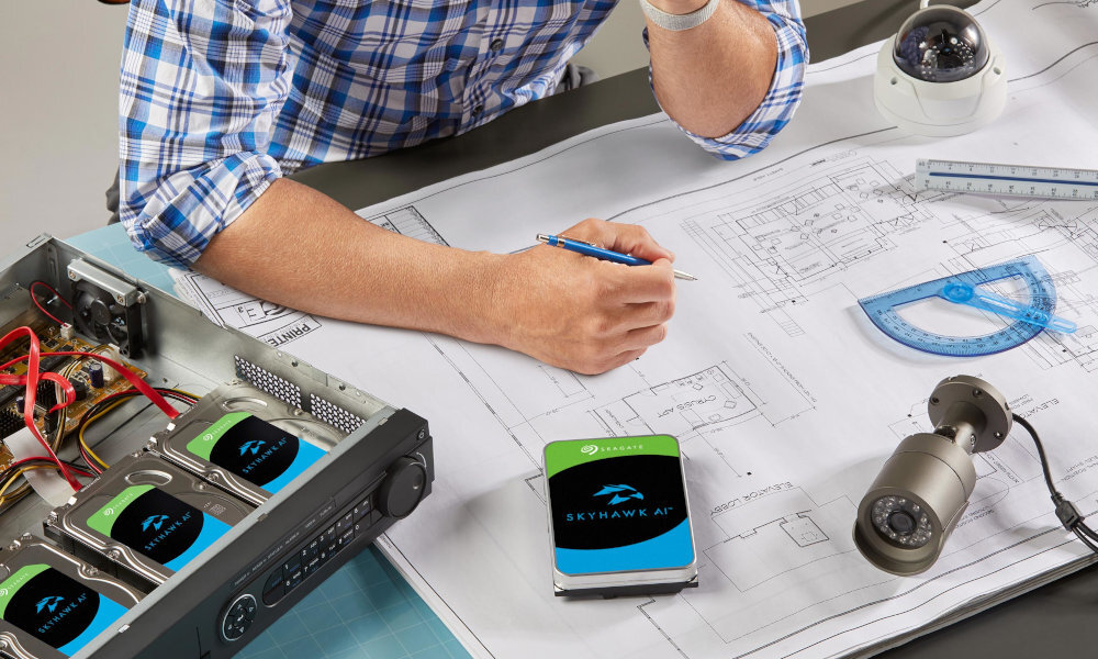 Na biurku leży dysk SEAGATE SkyHawk AI oraz kilka kamer i urządzeń elektronicznych. Osoba w koszuli w kratę rysuje na planach architektonicznych.