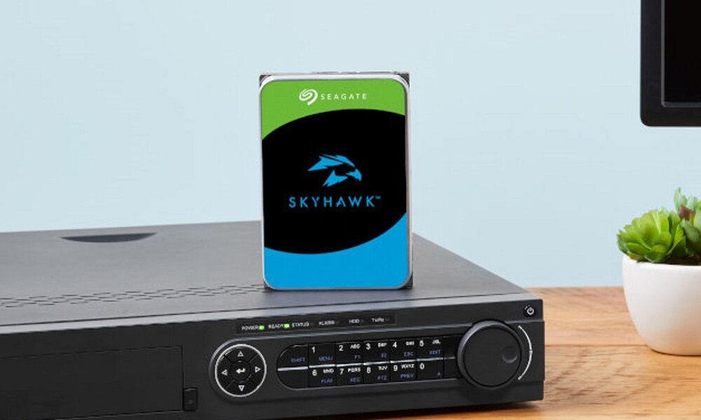 Na obrazie znajduje się dysk twardy z logo SEAGATE i napisem SKYHAWK, umieszczony na urządzeniu elektronicznym z przyciskami i wyświetlaczem. Obok znajduje się mała roślina w doniczce.