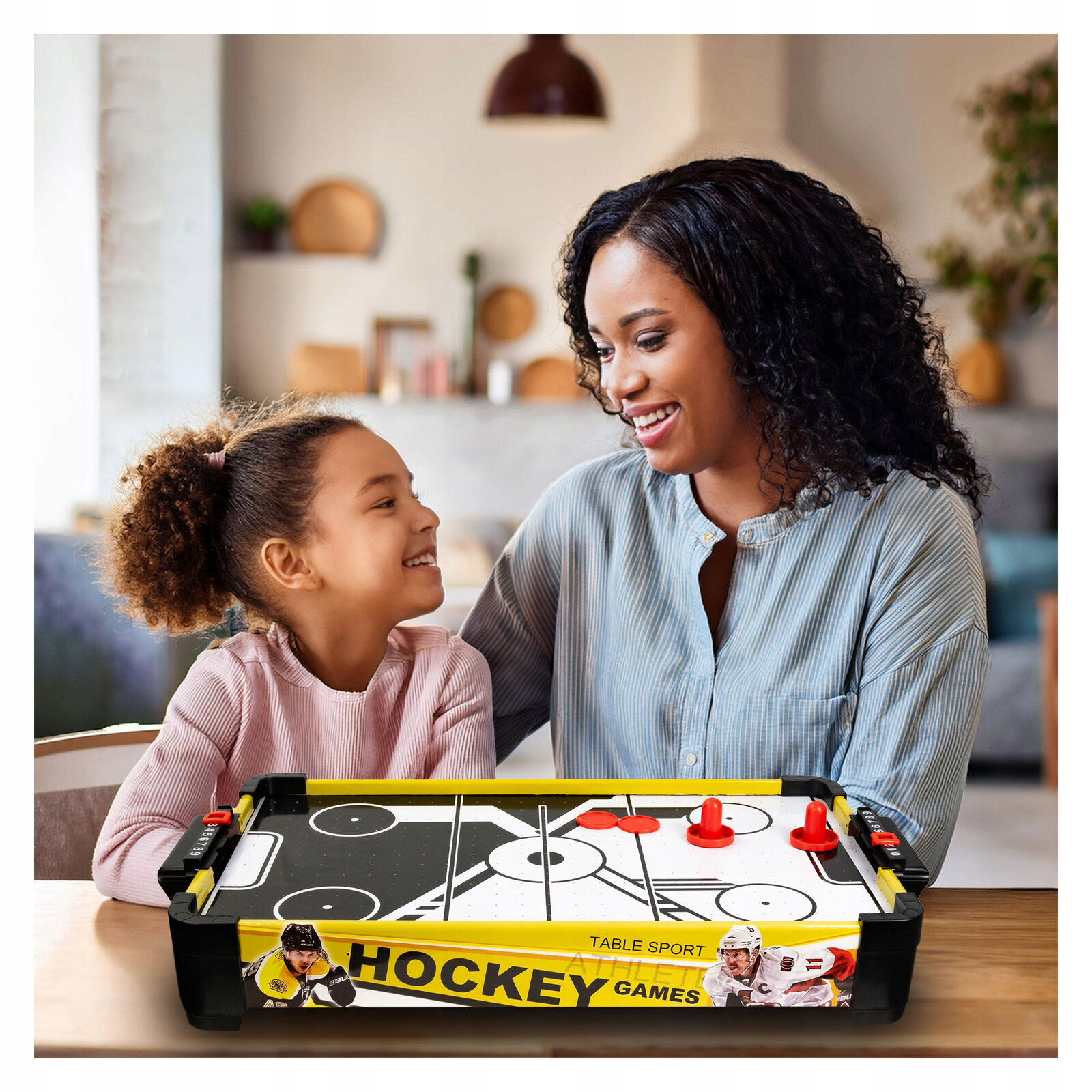 Na stole znajduje się stół do gry cymbergaj z systemem air flow, z widocznymi czerwonymi krążkami i odbijakami. Na boku stołu widnieje napis \'TABLE SPORT HOCKEY GAMES\'.