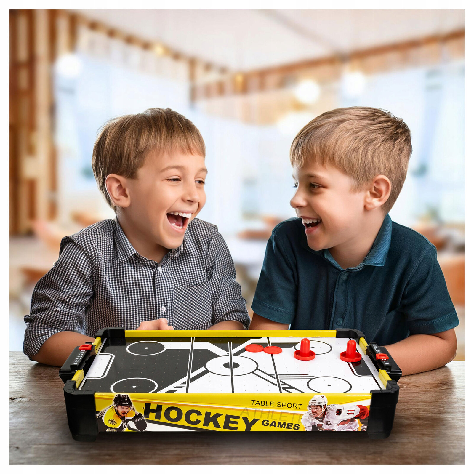 Dwóch chłopców siedzi przy stole do gry w cymbergaja z systemem air flow, na którym widoczne są czerwone krążki i odbijaki. Na boku stołu znajduje się napis \'HOCKEY GAMES\'.