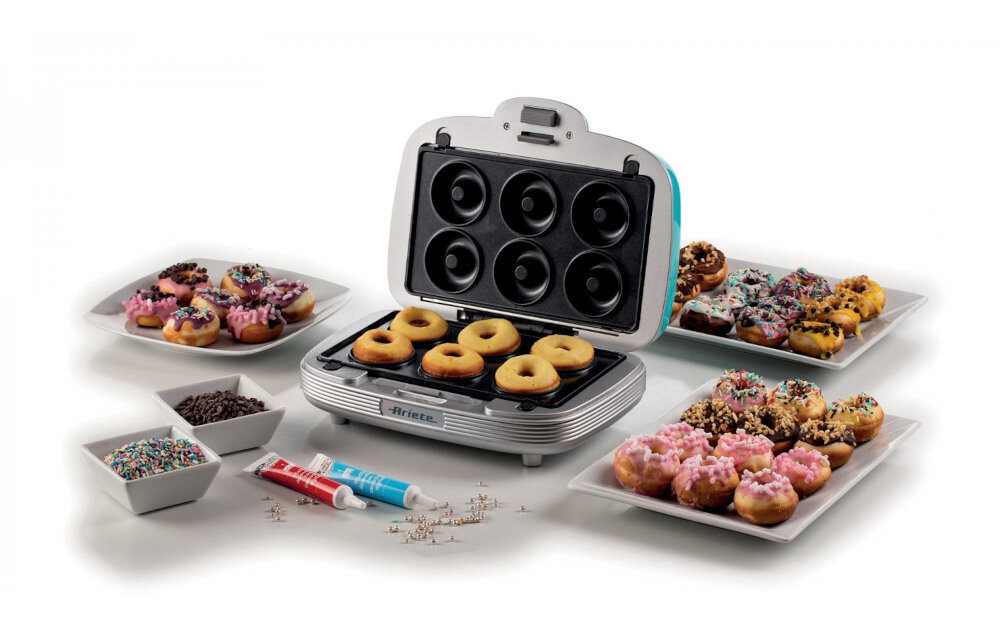 Opiekacz do donutów z sześcioma miejscami na ciasto, otwarty z gotowymi pączkami. Wokół znajdują się talerze z ozdobionymi donutami oraz miseczki z posypkami i tubki z lukrem.