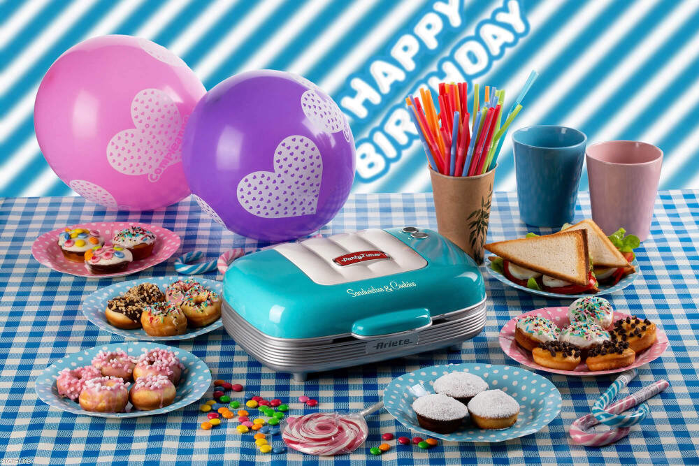 Na stole z niebiesko-białym obrusem znajduje się opiekacz Ariete w stylu retro, otoczony talerzami z kolorowymi ciastkami i kanapkami. W tle widoczne są balony z sercami oraz napis \'Happy Birthday\'.