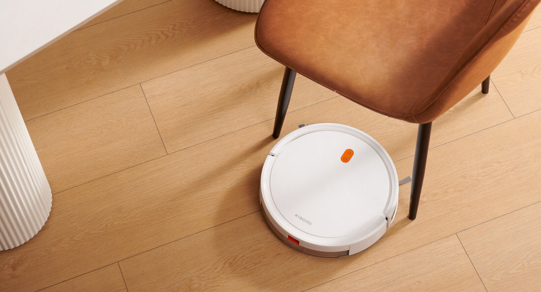 Biały robot sprzątający na drewnianej podłodze obok brązowego krzesła. Widoczny napis \'XIAOMI\' na urządzeniu.
